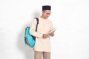 Portrait of excited student Asian muslim man in koko shirt with skullcap carrying backpack, while holding his school books. Islamic education concept. Isolated image on white background photo