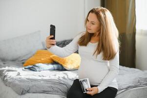 entonces bonito a oír usted hermosa joven embarazada mujer hablando en teléfono y mirando lejos con sonrisa mientras en pie a cocina interior foto