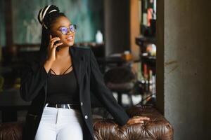 sonriente oscuro desollado mujer en de moda vestir lavando café hablando en móvil teléfono vía itinerancia aranceles, africano americano milenario hembra caminando y haciendo teléfono inteligente llamada satisfecho con conexión foto