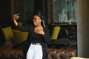 Cheerful dark skinned woman in trendy wear and spectacleson cafe posing for picture and media content for blog, smiling african american female 20s making selfie using mobile phone photo