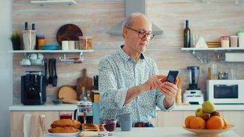 Senior man browsing on internet using smartphone in kitchen while enjoying morning coffee during breakfast. Authentic portrait of retired senior enjoying modern internet online technology video