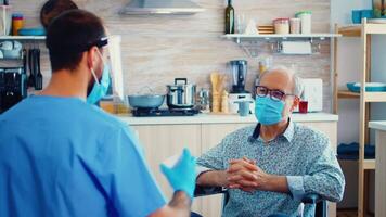 Disabled senior man in wheelchair discussing with caregiver about coronavirus during home visit. Social worker offering pills to handicapped elderly man. Geriatrician helping prevent covid-19 spread video