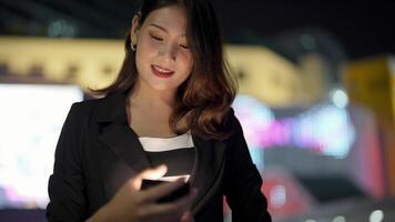 asiático mujer multirracial hermosa. negocio mujer utilizando teléfono inteligente en un ciudad calle con neón bokeh luces brillante a noche. mujer mirando a móvil teléfono caminando en calle con Encendiendo en cartelera. video