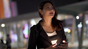 Aziatisch vrouw meerdere rassen mooi. bedrijf vrouw gebruik makend van smartphone Aan een stad straat met neon bokeh lichten schijnend Bij nacht. vrouw op zoek Bij mobiel telefoon wandelen in straat met verlichting Aan aanplakbord. video