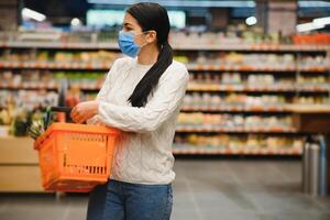 Alarmed female wears medical mask against coronavirus while grocery shopping in supermarket or store- health, safety and pandemic concept - young woman wearing protective mask and stockpiling food. photo