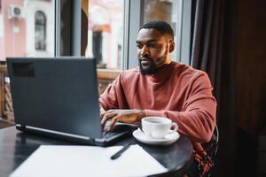 Portrait of thoughtful and concentrated young African American copy-writer, working on new articles using his laptop, and, looking at papers on a cafe table. photo