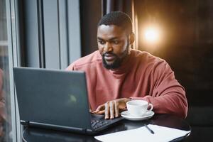 retrato de pensativo y concentrado joven africano americano redactor, trabajando en nuevo artículos utilizando su computadora portátil, y, mirando a documentos en un café mesa. foto