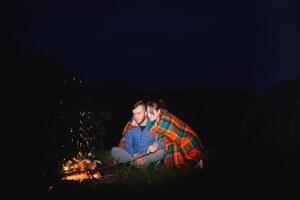 Young couple man and woman trevelers sitting near glowing tourist tent, burning campfire, on the top of mountain, enjoying beautiful view of night sky. photo