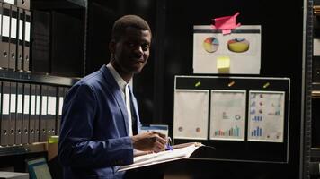 Determined black policeman standing and looking at camera in office full of evidence, files, and photos. Focused african american private investigator holding and reviewing crimanal records. Portait. photo