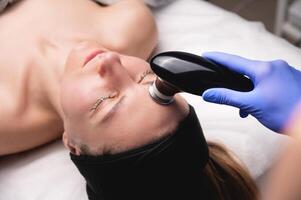 Young woman undergoing facial rejuvenation procedure with tekar therapy in salon, top side view, close-up photo