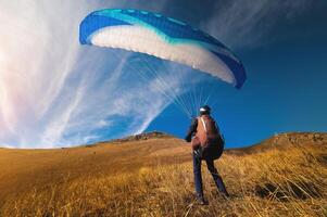 paraglider stands on yellow grass in a field and holds his parachute in the air. Extreme sport photo