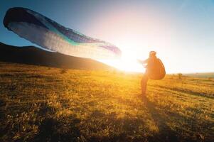 paraglider with a blue parachute takes off. A male athlete stands on the field and lifts a paraglider into the air photo