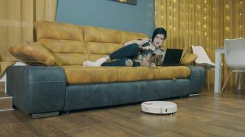 A woman lying on a sofa and working on a laptop, and a robot vacuum cleaner automatically vacuumes the room. Automated room cleaning. Free time to work with your pet. video