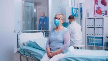 retrato do idosos senhora sentado em a hospital cama durante covid-19 crise. médico e pacientes vestindo protetora mascarar e equipamento dentro moderno privado cínico. cuidados de saúde sistema depois de coronavírus video