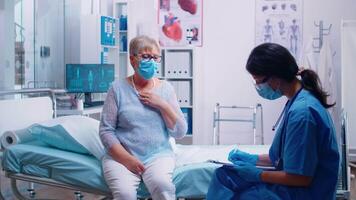 Female nurse talking with old senior patient, writing down answears. COVID 19 pandemic medical consultation. Modern private clinic or hospital. Practitioner physician appointment video