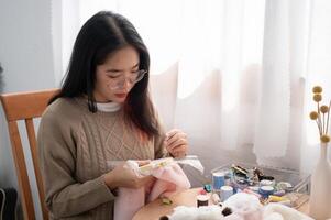 un enfocado joven asiático mujer enhebrar un modelo en un bordado marco, coser a mano en paño. foto