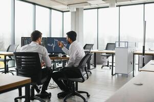 Two men traders sitting at desk at office together monitoring stocks data candle charts on screen analyzing price flow smiling cheerful having profit teamwork concept photo