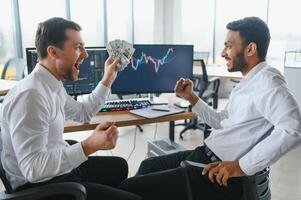 Two men traders sitting at desk at office together monitoring stocks data candle charts on screen analyzing price flow smiling cheerful having profit teamwork concept photo