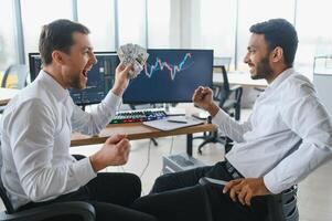 dos hombres comerciantes sentado a escritorio a oficina juntos supervisión cepo datos vela cartas en pantalla analizando precio fluir sonriente alegre teniendo lucro trabajo en equipo concepto foto