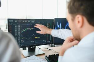 Two men traders sitting at desk at office together monitoring stocks data candle charts on screen analyzing price flow smiling cheerful having profit teamwork concept photo