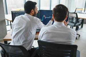 Two men traders sitting at desk at office together monitoring stocks data candle charts on screen analyzing price flow smiling cheerful having profit teamwork concept photo