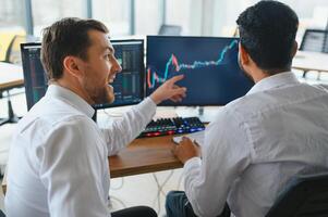 Two men traders sitting at desk at office together monitoring stocks data candle charts on screen analyzing price flow smiling cheerful having profit teamwork concept photo