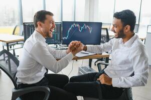 Two men traders sitting at desk at office together monitoring stocks data candle charts on screen analyzing price flow smiling cheerful having profit teamwork concept photo