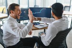 Two men traders sitting at desk at office together monitoring stocks data candle charts on screen analyzing price flow smiling cheerful having profit teamwork concept photo