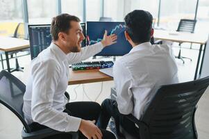 Two men traders sitting at desk at office together monitoring stocks data candle charts on screen analyzing price flow smiling cheerful having profit teamwork concept photo