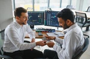 Two men traders sitting at desk at office together monitoring stocks data candle charts on screen analyzing price flow smiling cheerful having profit teamwork concept photo