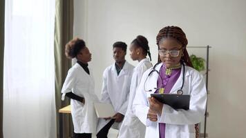 africano americano mulher médico dentro branco vestido e óculos olhando às Câmera e sorridente dentro clínica. médicos em fundo video