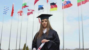 A university graduate stands on the background of flags of the world holding graduatind diploma of Master's degree of international standard. View of the open space. national flags in the blue sky video