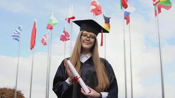 A university graduate stands on the background of flags of the world holding graduatind diploma of Master's degree in her hands. View of the open space. National flags in the blue sky video