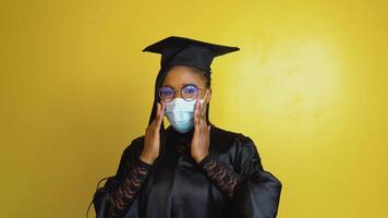 The graduate student in a protective mask expresses with gestures the need to comply with quarantine security measures. Student in a black robe and hat on a yellow solid background looks at the camera video