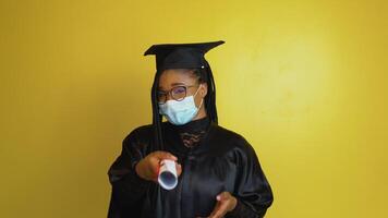 A graduate student in a protective mask points with a diploma in hand towards the camera. Student in a black robe and hat on a yellow solid background looks at the camera video