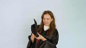 Young caucasian female student in a black robe lightly throws a square hat with a tassel up and catches.White background video