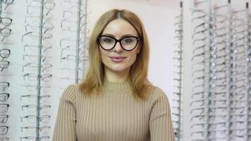 attrayant Jeune blond femme permanent dans de face de une miroir portant transparent des lunettes avec une noir Cadre pour vision correction dans un lunettes boutique et à la recherche à le caméravision correction concept video