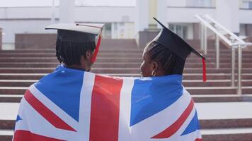 African American man and woman standing side by side with their backs to the camera.They have the British flag on their shoulders. A University building on background video