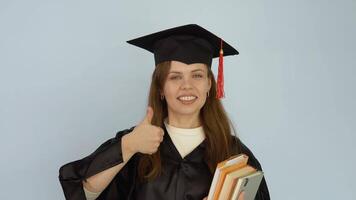 A young Caucasian woman in a black robe and a master's hat stands straight holding textbooks and showing agesture good with big finger up. Student looks at the camera on white background video