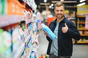 Young cheerful positive male customer making purchases in supermarket, buying household chemicals photo