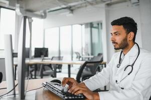 Indian male doctor physician wearing glasses, white medical gown and stethoscope sitting at the desk with the laptop in modern clinic and involved online video onference, consulting remotely. photo