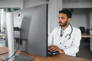 indio masculino médico médico vistiendo anteojos, blanco médico vestido y estetoscopio sentado a el escritorio con el ordenador portátil en moderno clínica y involucrado en línea vídeo conferencia, consultante remotamente foto