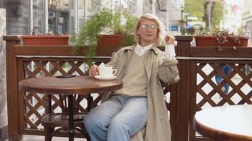 vrouw koude rillingen uit terwijl resting zittend met een kop van koffie Bij een tafel Aan de terras. een vrouw in een loopgraaf jas en wit blouse picks omhoog een wit koffie kop en zet het terug Aan de tafel video