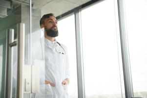 Young 30-aged handsome pleasant Arabic male doctor in white coat, posing at camera indoors. photo