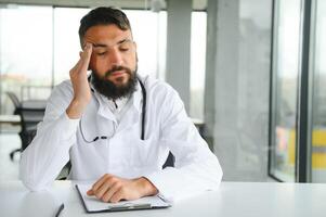 Young 30-aged handsome pleasant Arabic male doctor in white coat, posing at camera indoors. photo