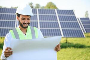 retrato de joven indio hombre técnico vistiendo blanco difícil sombrero en pie cerca solar paneles en contra azul cielo.industrial trabajador solar sistema instalación, renovable verde energía Generacion concepto. foto