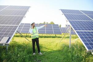 Portrait of Young indian man technician wearing white hard hat standing near solar panels against blue sky.Industrial worker solar system installation, renewable green energy generation concept. photo