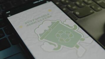 Sustainable agriculture inscription on smartphone screen. Green friendly plastic cup image with a recycle sign on grey background. Environment concept. Male hand flapping fingers cheerfully video