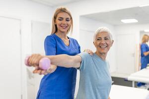 Oder aged woman doing physiotherapy with support from physiotherapists . Senior elderly female sitting in clinic using dumbbells workout exercise for patient with caregiver in nursing care. photo