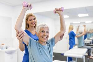 Oder aged woman doing physiotherapy with support from physiotherapists . Senior elderly female sitting in clinic using dumbbells workout exercise for patient with caregiver in nursing care. photo